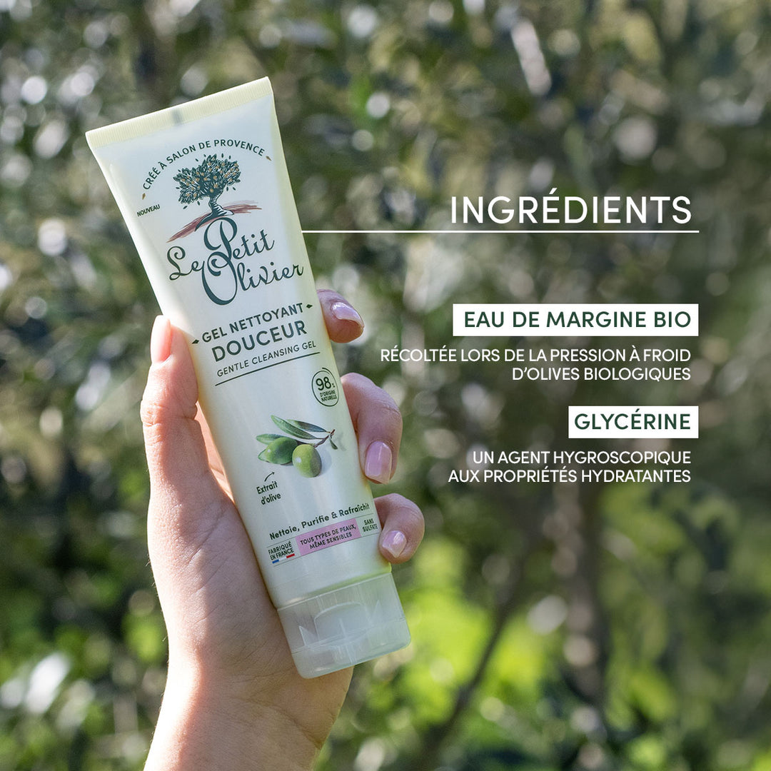 À l'extérieur, devant un décor végétal, une main tient un tube de gel nettoyant visage « Douceur » de Le Petit Olivier. L'étiquette met en avant l'huile d'olive bio et la glycérine, et décrit ce nettoyant visage sans sulfate comme hydratant et doux pour les peaux sensibles.