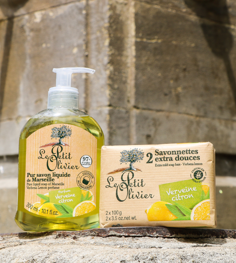 Du savon liquide Le Petit Olivier et deux pains de savon extra-doux, tous deux au parfum Verveine Citron, présentés à l'extérieur sur une surface en pierre, avec un arrière-plan flou.