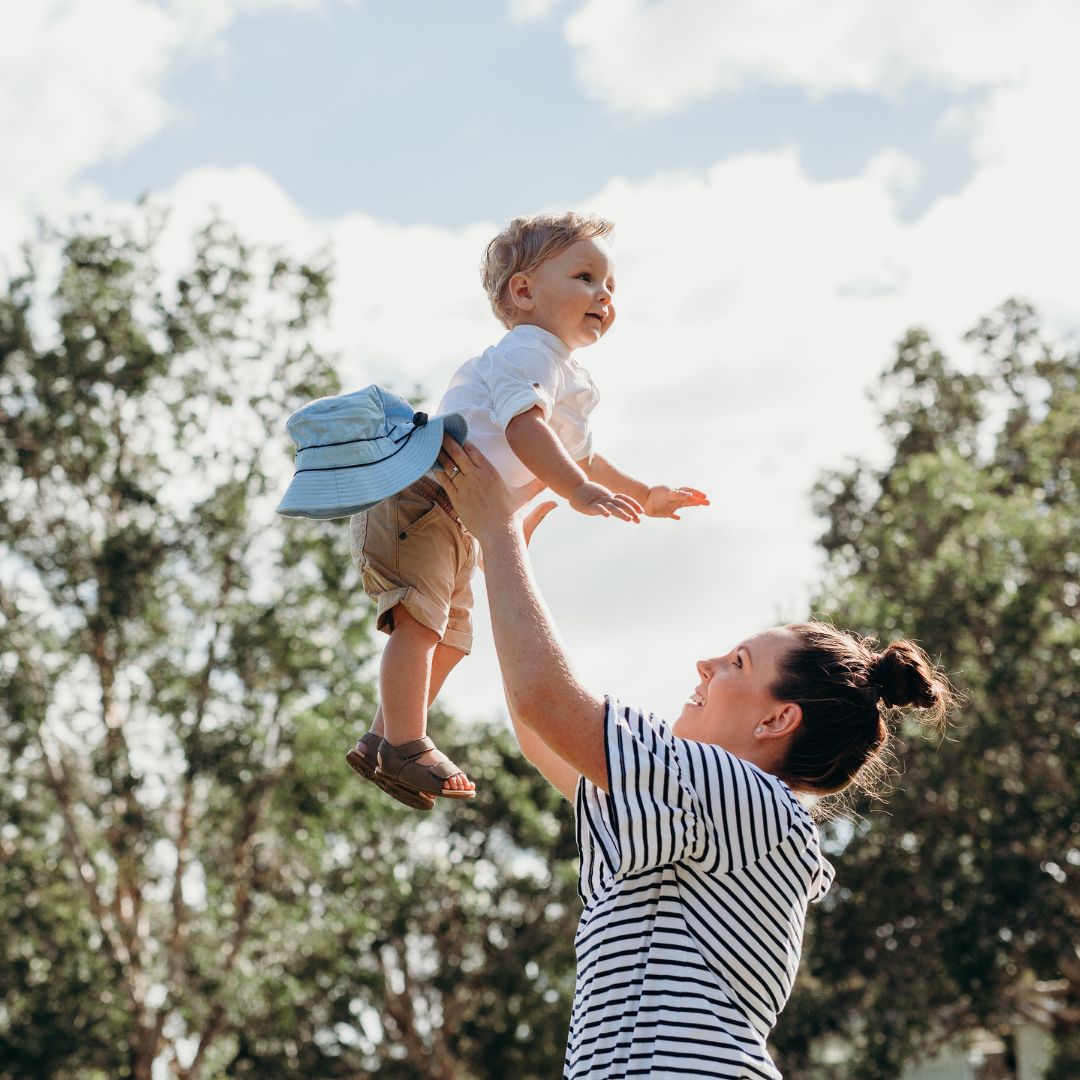 Une femme vêtue d'un t-shirt rayé soulève dans les airs un petit enfant souriant, à l'extérieur. Le ciel est partiellement nuageux et on aperçoit des arbres à l'arrière-plan.