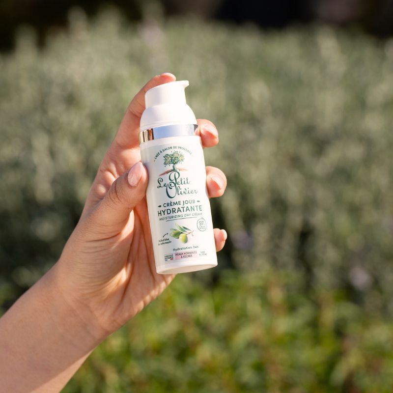 Une main tient un flacon de crème hydratante « Le Petit Olivier Crème Riche Hydratante » à l'extérieur, sur un fond flou aux tons verts et gris.