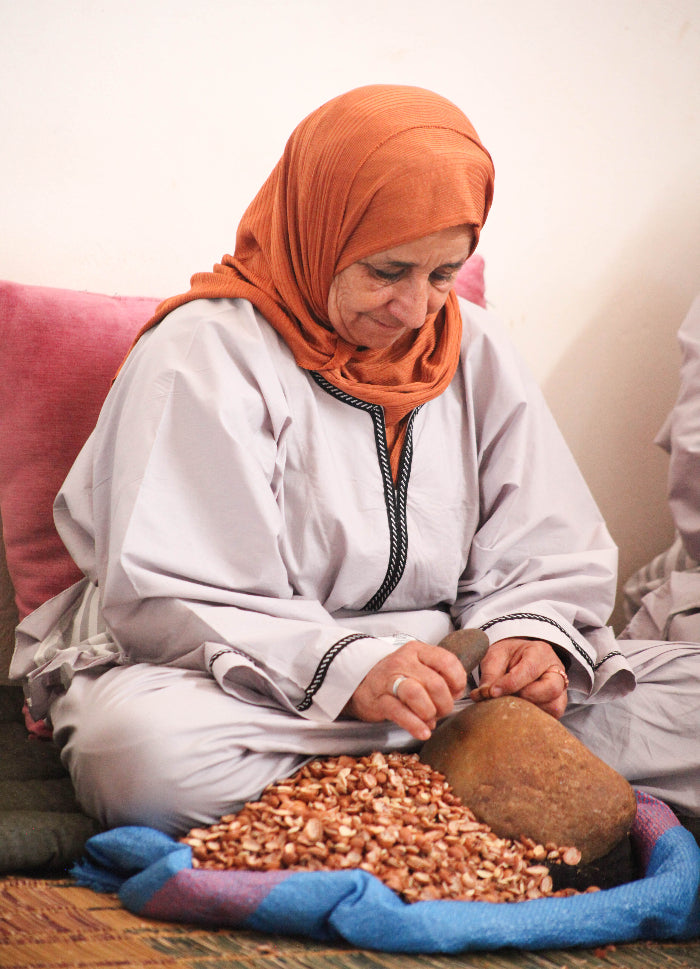 Une femme vêtue d'une robe légère et d'un foulard orange est assise en tailleur ; elle casse des noix à l'aide d'un outil posé sur une pierre, tandis qu'un tas de noix est disposé sur un tissu bleu devant elle.