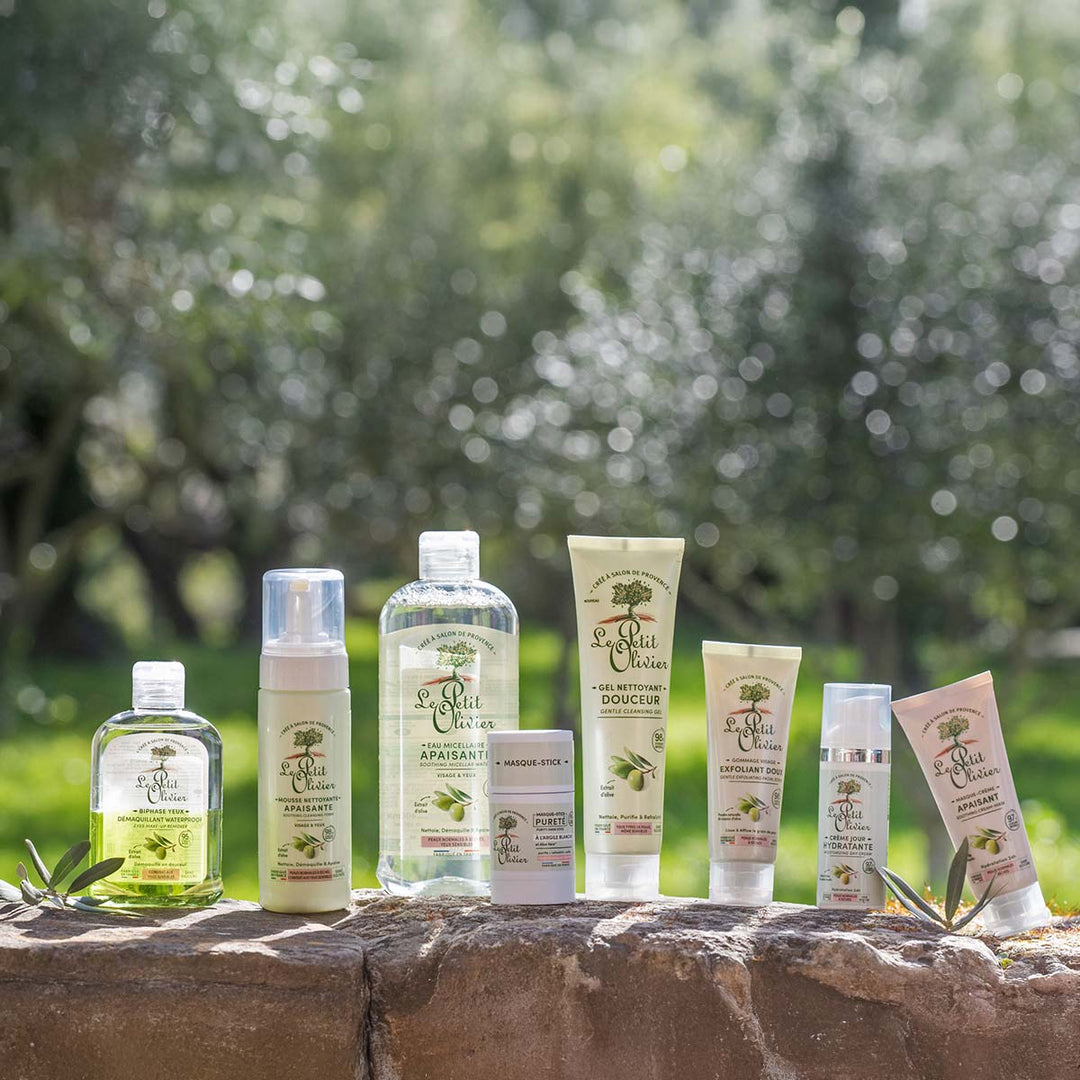 Le Gel Nettoyant Visage Douceur de Le Petit Olivier, ainsi que cinq autres produits de soin, sont présentés en extérieur sur un rebord en pierre, avec en arrière-plan une vue floue d'arbres verts et d'herbe, évoquant un cadre naturel.